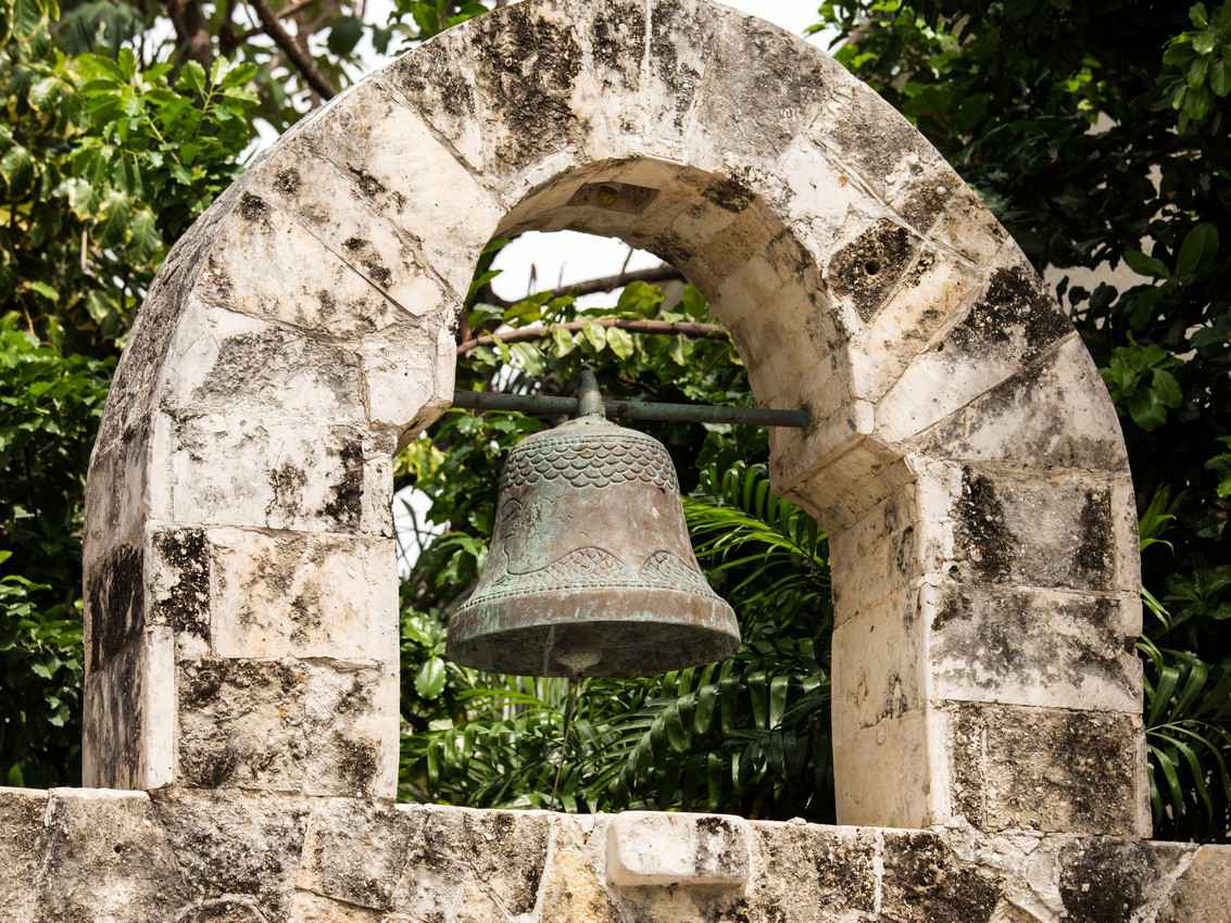 An old and antiquated bell that is hanging from a stone arch in downtown Playa Del Carmen.