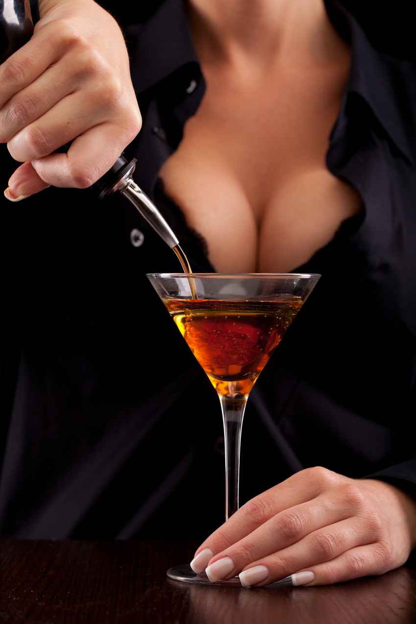 A sexy bartender with big boobs pouring a drink into a martini glass.