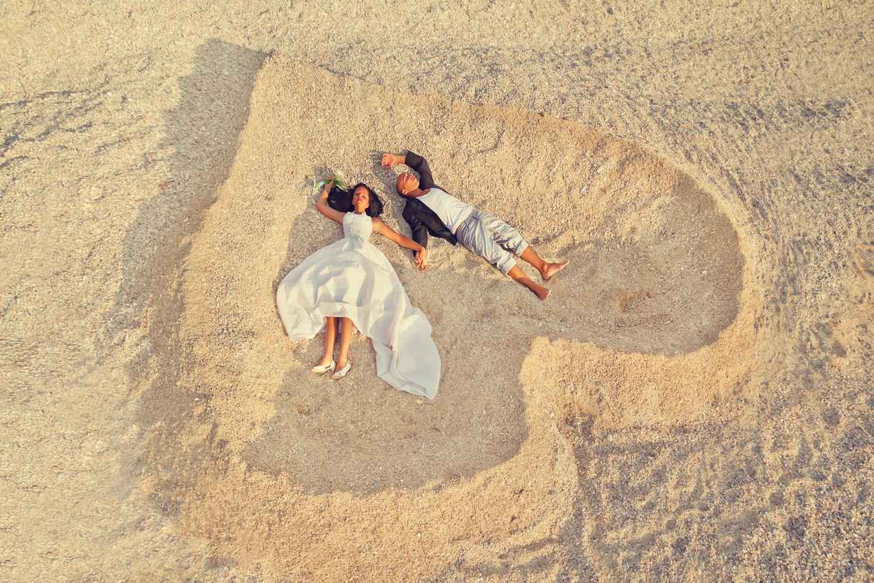 A man and a woman lying on the beach making a heart in the sand.