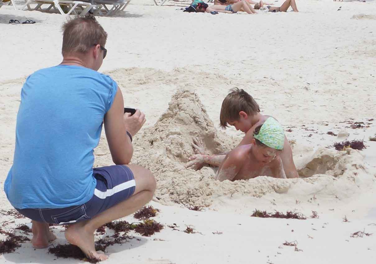 A man on the beach taking photographs of his children who are digging a hole in the sand.