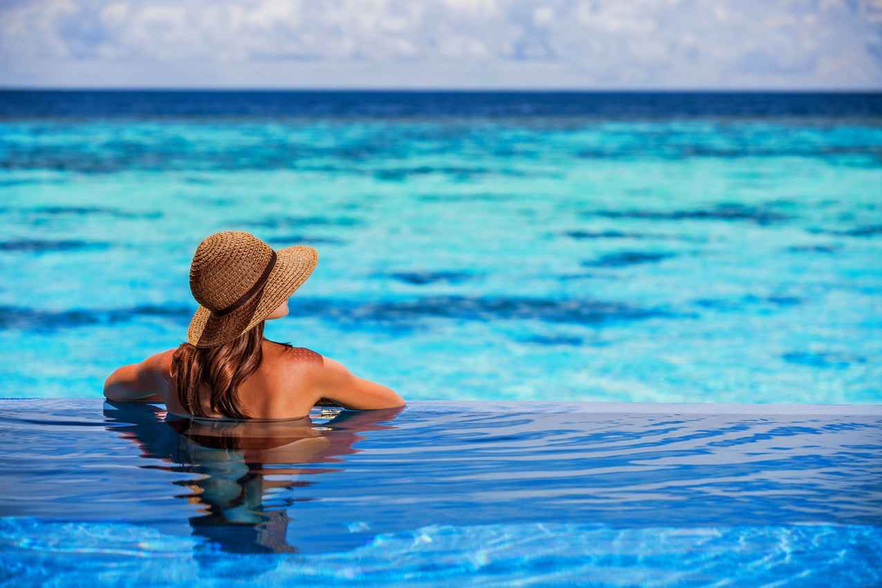 A woman wearing a sun hat in an infinity pool overlooking the crystal clear blue water.