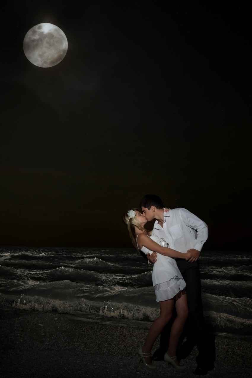 A man and a woman kissing on the beach with a full moon in the background.