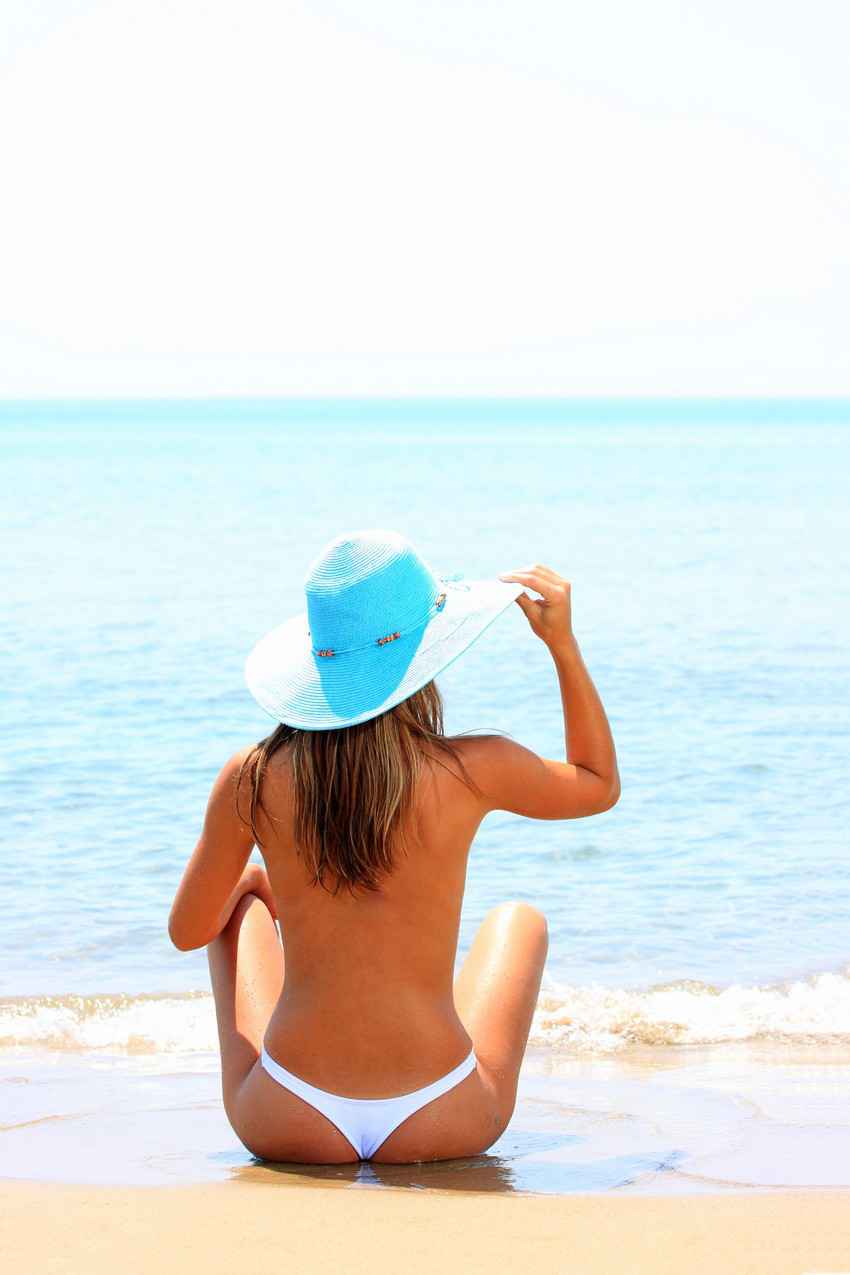 A topless woman on the beach wearing a white bikini and a blue hat.