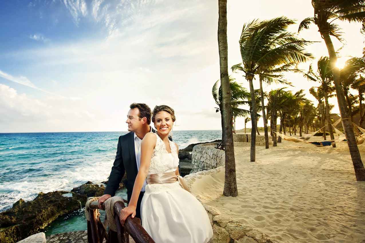 A recently married man and woman standing near the beach with palm trees and hammocks behind them.