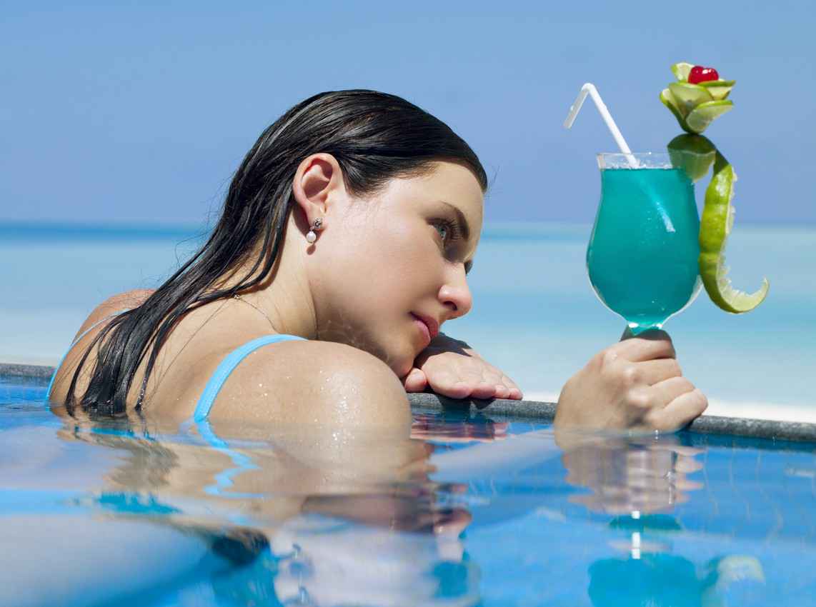 A beautiful woman in a swimming pool overlooking the beach holding a cocktail in her hand.