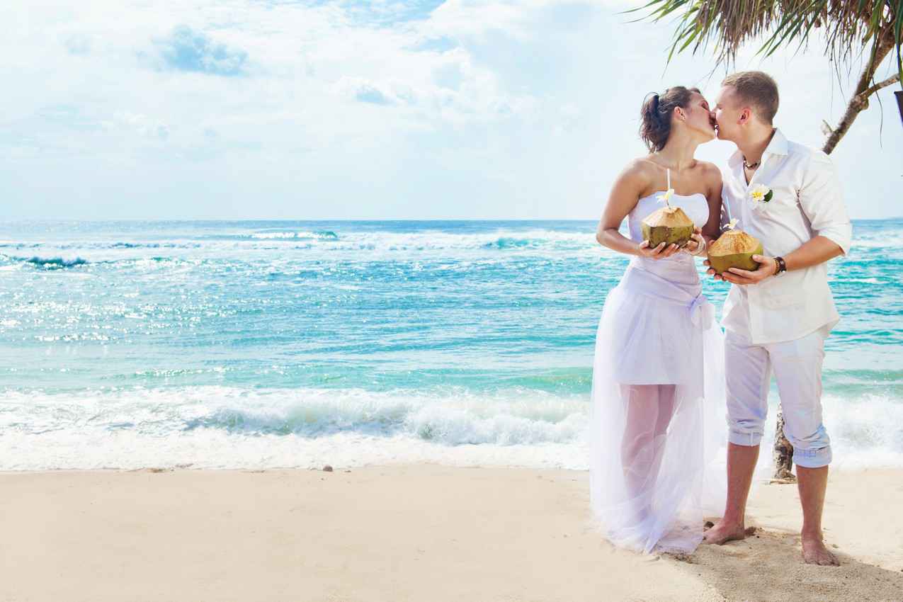 A bride and groom kissing on the beach holding several coconut drinks.