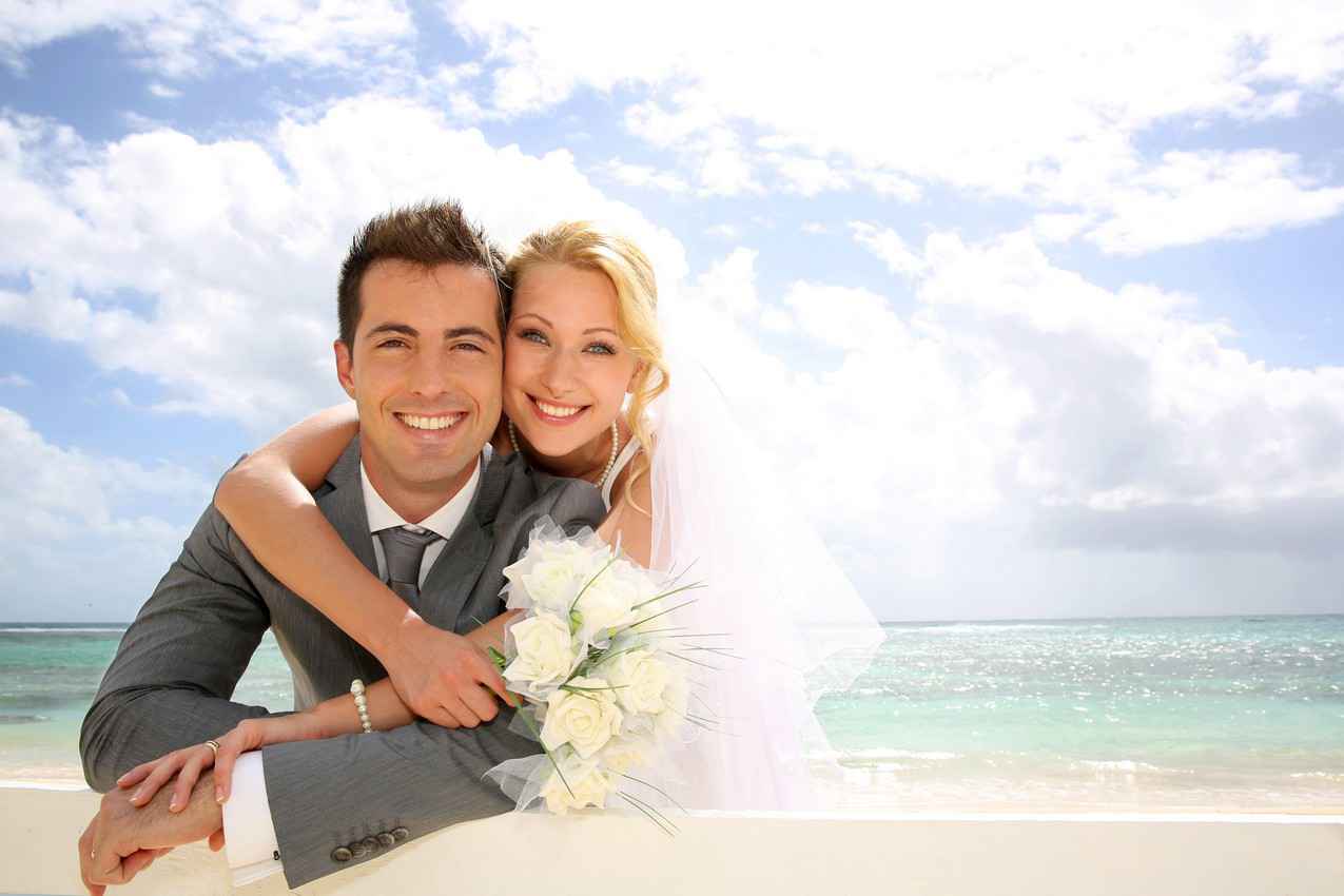A bride and groom on the beach with bright sun, white sand, and beautiful blue water visible behind them.