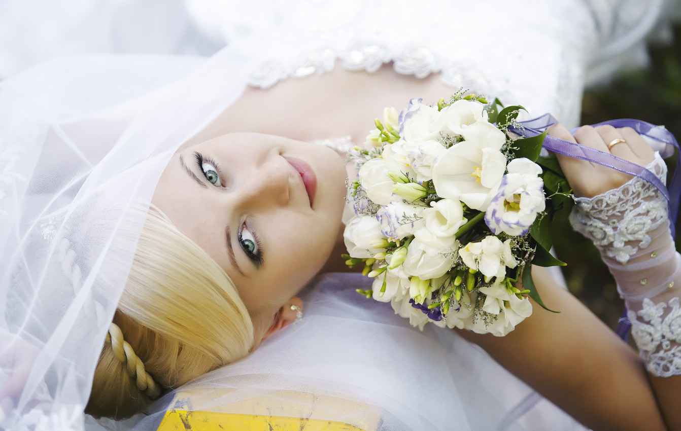 A bride laying down on a bench with a flower bouquet and very beautiful hair.