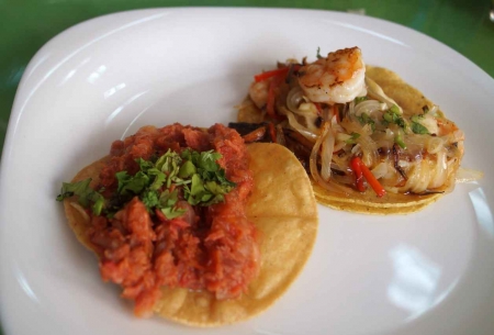 A pork taco next to a seafood taco with both served on soft corn tortillas.