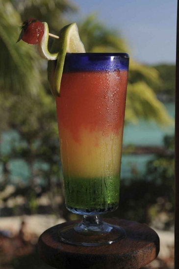 A multicolored Mexican cocktail with palm trees and water in the background.