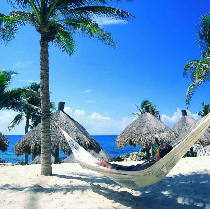 A woman lying in a beech hammock with several palapas behind her.