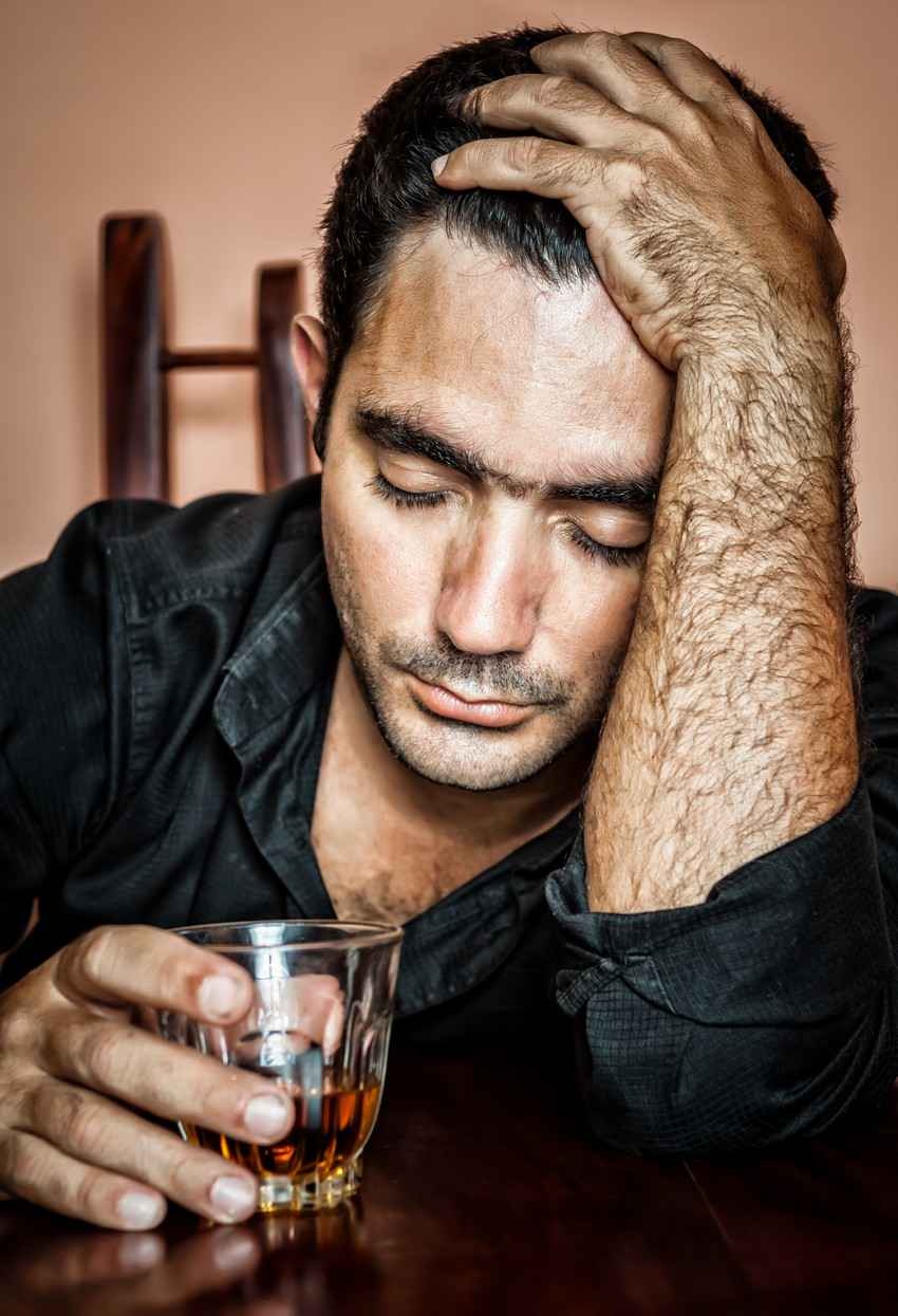 A drunk hairy man falling asleep at a bar with a whiskey glass in his hand.