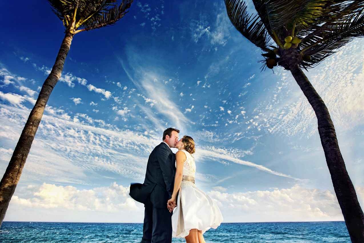 A bride and a groom on the beach with the sea on one side and palm trees on the other side.