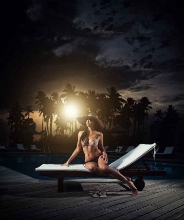 A sexy brunette sitting on a lounge chair in front of a pool with the sunset in the background.