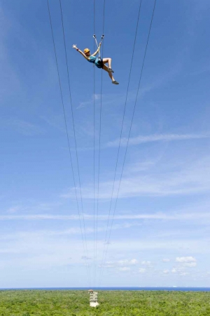 A woman wearing a yellow helmet who is high above the jungle canopy on a zip line.