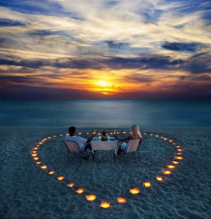A recently married couple sitting on the beach watching the sunset inside of a large heart that was created with burning candles.