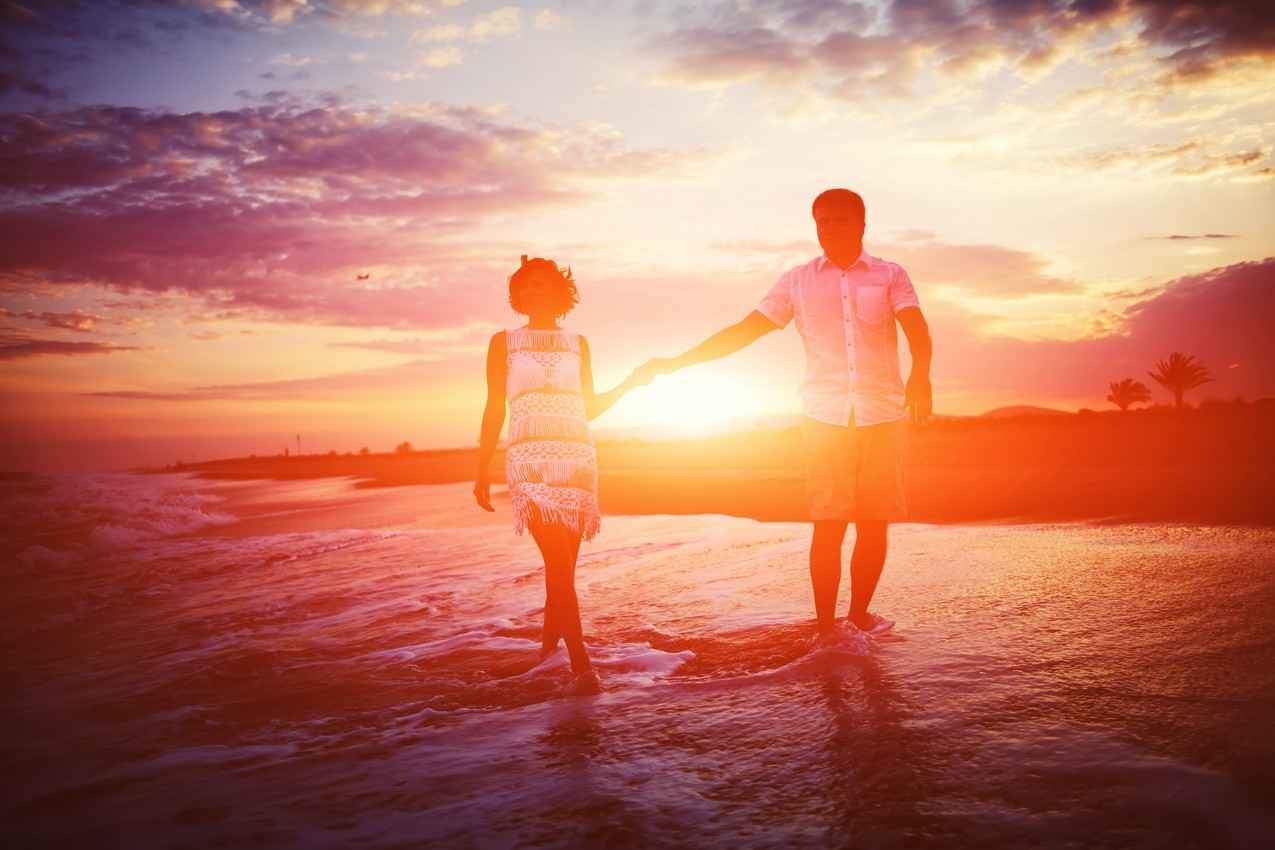 A man and a woman walking on the beach at sunset with waves lapping at their feet.
