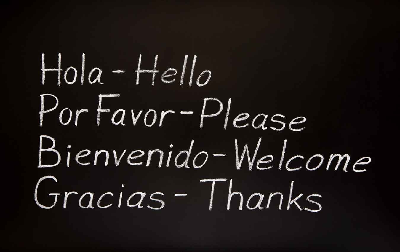 The words, hello, please, welcome, and thanks translated from Spanish to English and written on a black chalkboard.