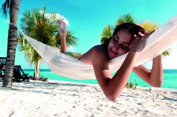 A woman is lying in a hammock with white sand and aqua blue water behind her.